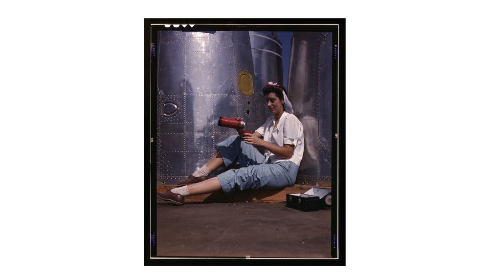 A young woman sits against a shining silver tank, pouring from a thermos into a cup. an open lunch pail is next to her. 