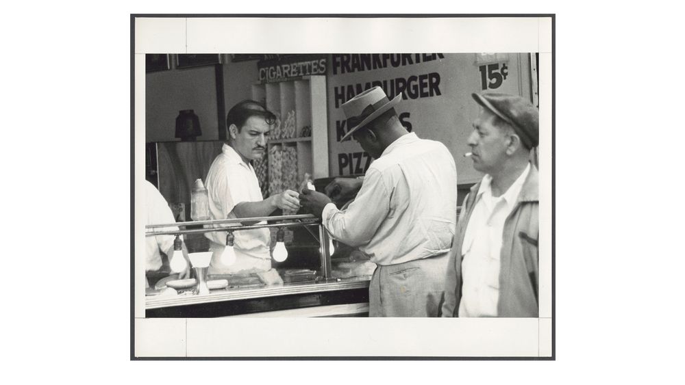 b&w photograph showing a man at a lunch counter serving another man with his back to the viewer. The sign behind the stand reads Frankfurter, Hamburger 15c, Pizza. A storage area next to the sign holds just a shit ton of cigarettes. At the right, a man with a stubby cigarette walks past while glancing by. 