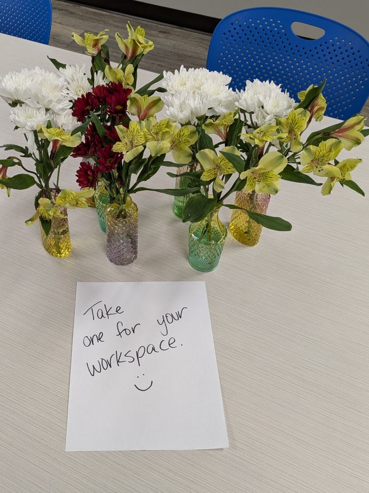 Several small vases of flowers on a lunchroom table with a note that says "take one for your workspace"