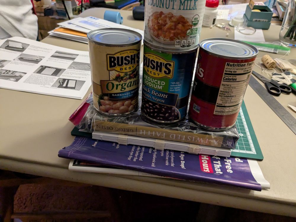 a stack of book signatures on top of scrap paper, beneath a book and several cans of food weighing it all down as glue dries on the spine