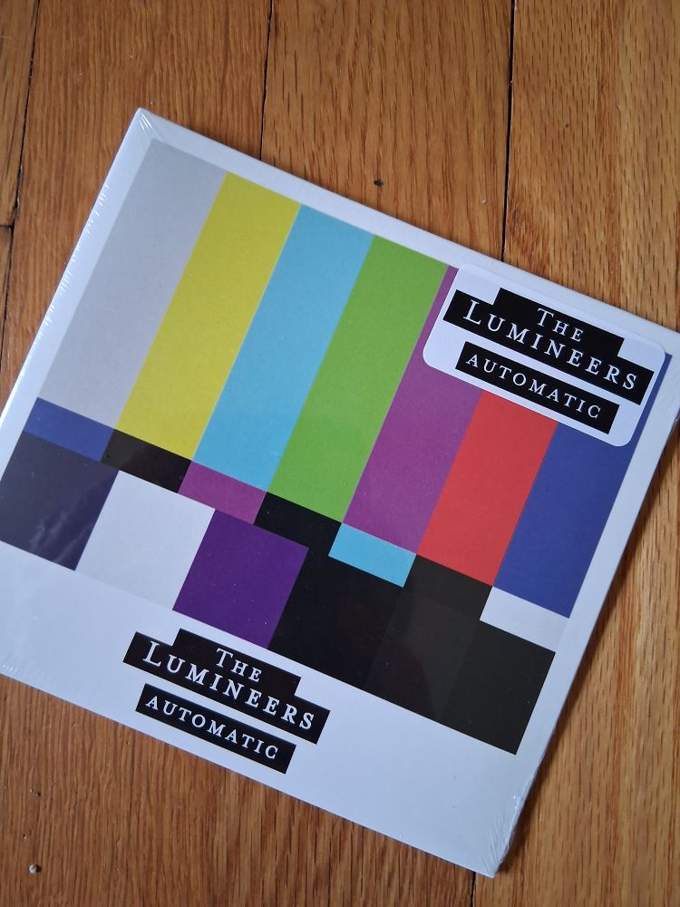 A still wrapped cd. It sits on a wooden floor. It is automatic by the lumineers. It is a white cover with the rainbow bar thing that used to appear on old TV screens when TV shows stopped for the day