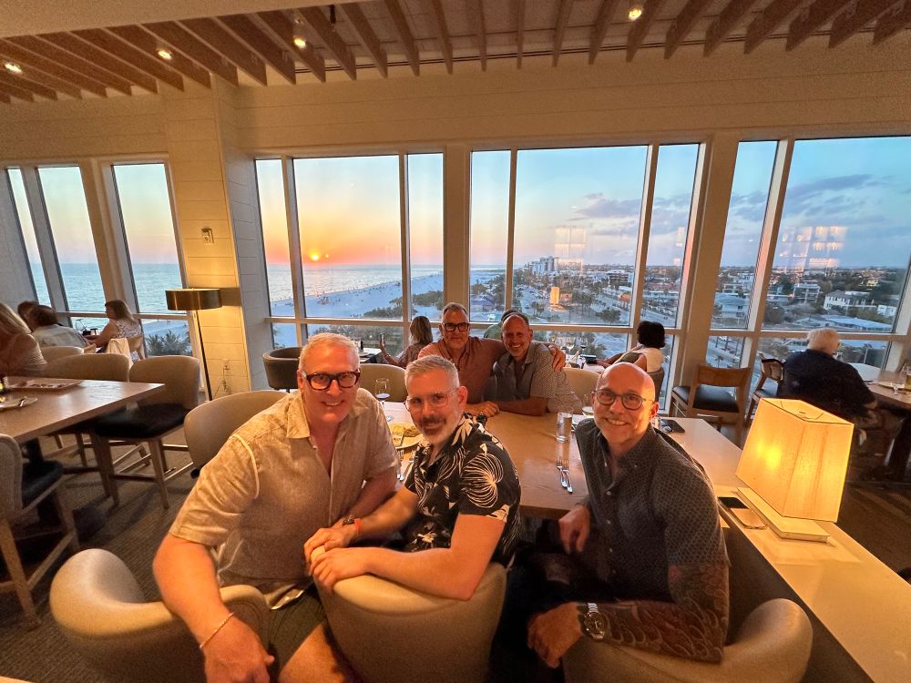 A picture of five aging but still young white gay guys sitting in a hotel restaurant with windows behind us showing the gulf coast FL sunset in the background. 
