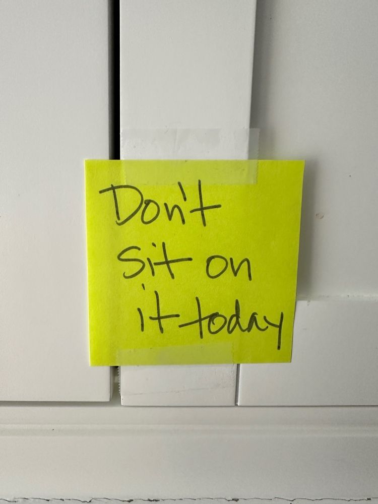 A yellow post-it note on a white bathroom cabinet reading "Don't Sit on It Today."