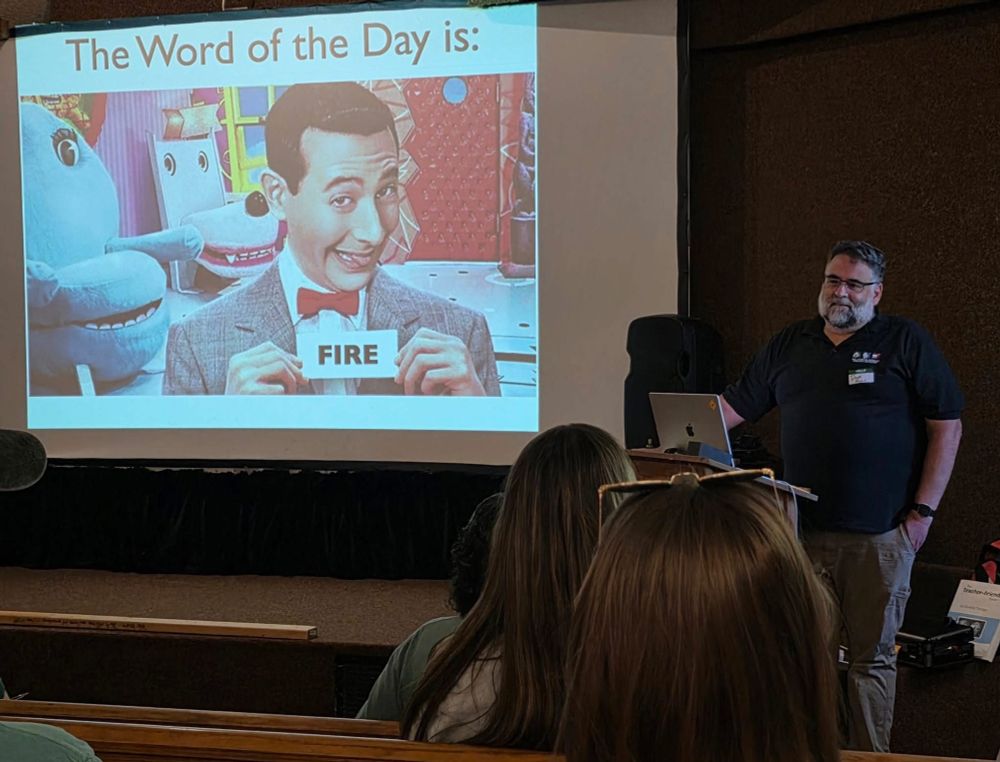 Don in front of a projected image of Peewee Herman holding up a card that simply reads, “ FIRE.” Above the image, text says, “The Word of the Day is:”