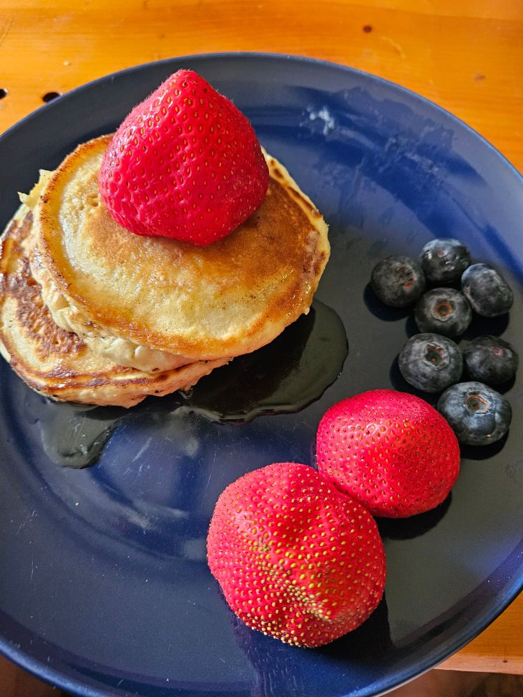 Three pancakes, topped with syrup, accompanied by 3 strawberries and 7 blueberries. 