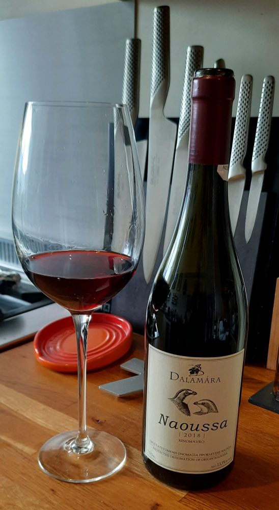 Bottle of red Greek wine next to part filled large wine glass on kitchen top.