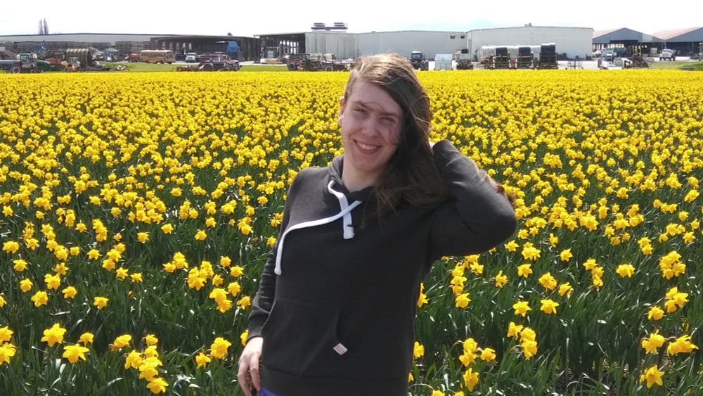 photo of a young snacks, a white nonbinary trans woman, standing in front of a large field of bright yellow daffodils. she’s wearing a grey hoodie and the bright white drawstrings of her hood rest on the small outline of her breasts. she’s raising one arm up behind her head as the wind blows her shoulder length (😱) brown hair forward. she’s smiling brightly at the camera