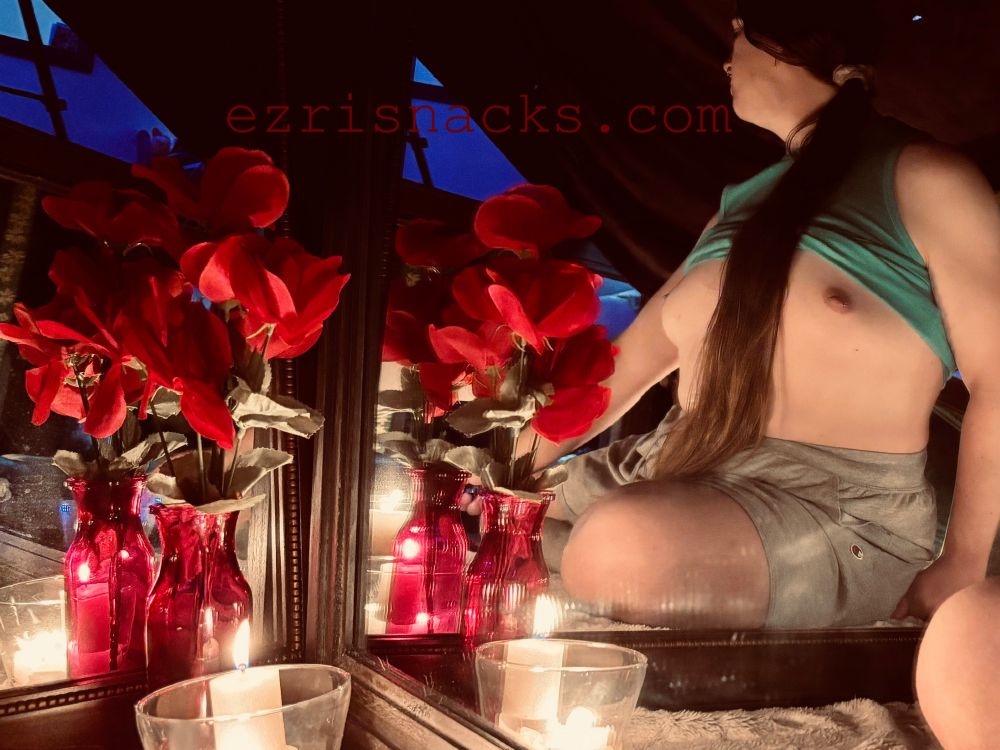 photo of snacks, a white non binary trans woman, she’s wearing a green tank top and grey shorts and facing away from the camera, her tank top is pulled up to unveil her tiny trans titties and pierced nipples. her long smooth shiny brown hair is tied in a ponytail and flows down the full length of her torso. in the foreground is a pink glass vase of red roses and a clear glass candle holder, in which are three lit white candles. above the roses is red text that reads ezrisnacks.com 