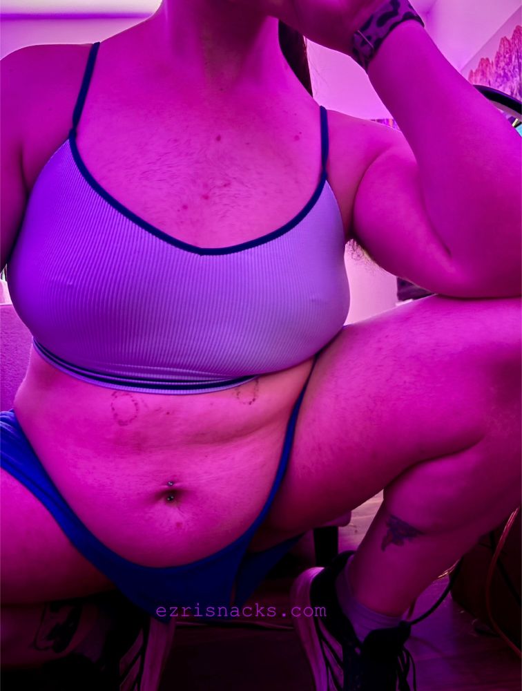 photo of snacks, a white non binary trans woman, crouched down, balanced on her tip toes, and posed with her legs open, belly out, and left arm perched on her left knee. she’s wearing blue shorts and a light blue bralette, black adidas shoes, and her face is out of frame. text on the image reads ezrisnacks.com 
