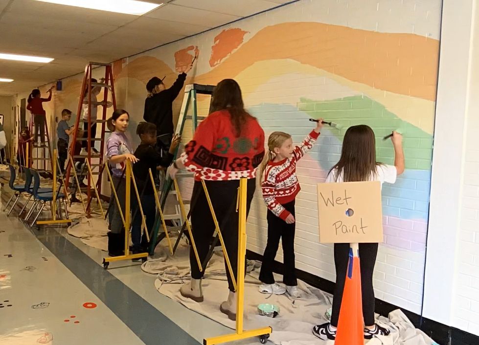 Students in a school hallway painting a rainbow on the wall. Some are on ladders, some are standing on the ground. 