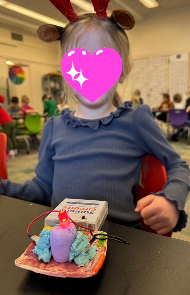 Young preschool girl wearing reindeer antlers sits at a table in front of a project. She has a clay votive, play doh and clay hooked up to an LED Light and a squishy circuit  