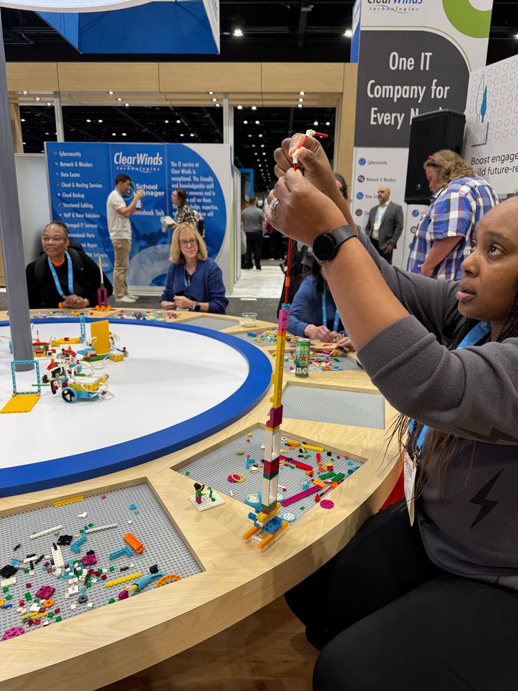 Teachers sitting around a round LEGO table engaged in building tall structures out of LEGO bricks