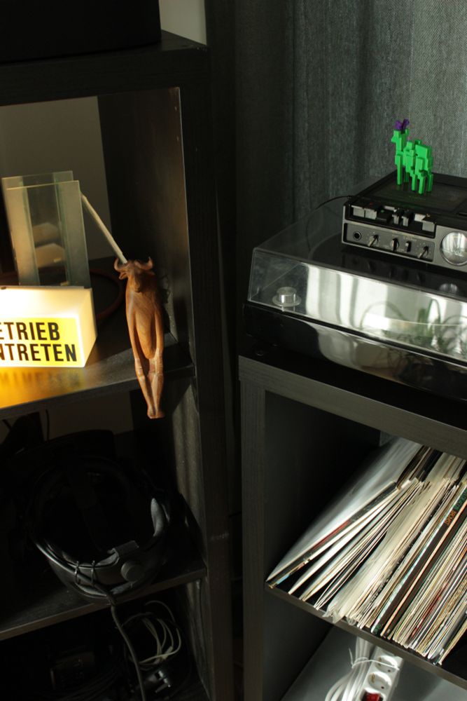 My shelves with knickknacks and vinyls and record player and tape player. The tone is dark greens and blacks, with the knickknacks contrasting in glowing yellow (lamp), warm brown, neon green.