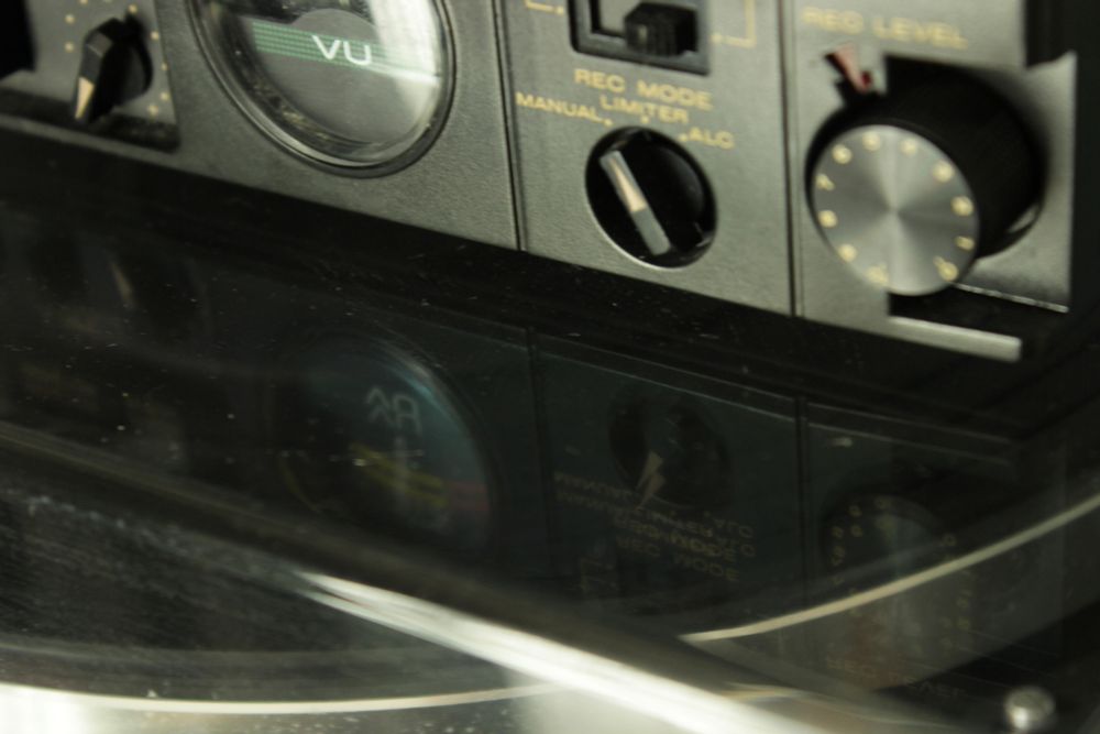 Getting closer to the tape player on top of the record players clear lid, looking at the reflection of the tape player, showing it's VU meter, compressor and level settings.
