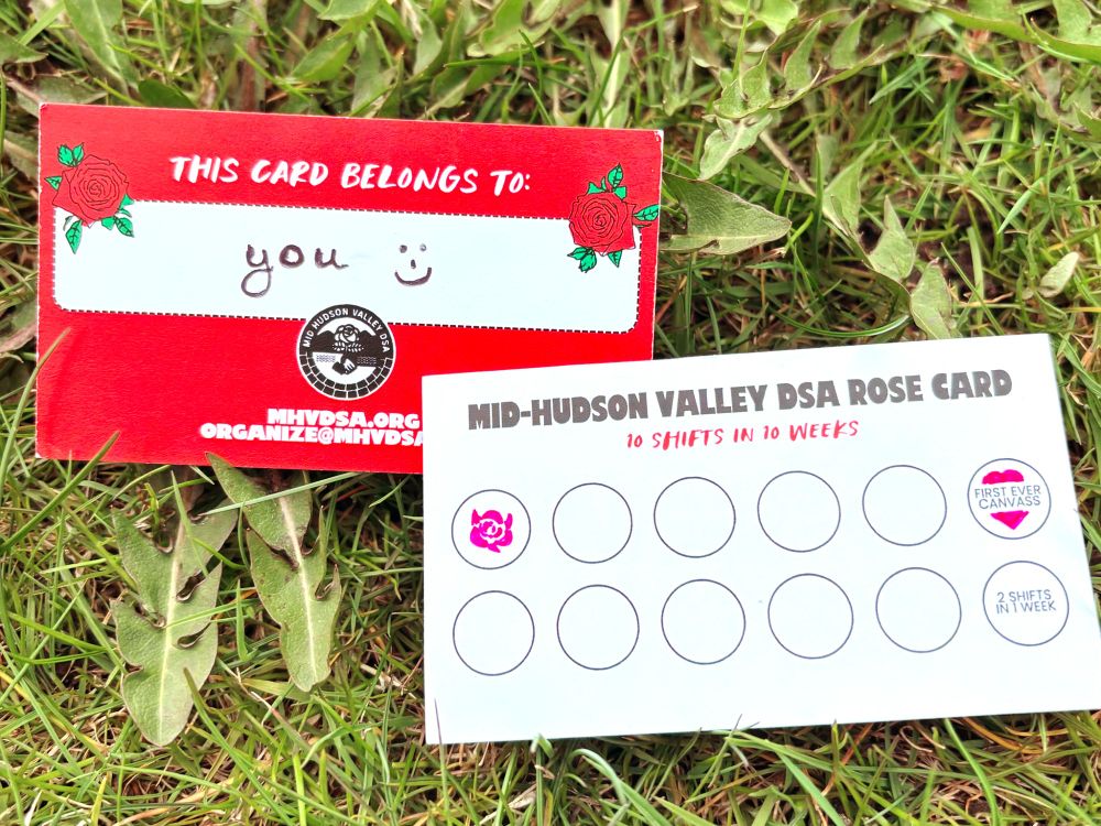 The image shows two cards placed on green grass. The card on the left is predominantly red with a decorative rose in each corner. It has handwritten text in a central white box reading "you :)" and includes logos and text at the bottom related to the Mid-Hudson Valley DSA. The card on the right is mostly white with "MID-HUDSON VALLEY DSA ROSE CARD" at the top, followed by "10 SHIFTS IN 10 WEEKS" in red. Below are ten empty circles, two of which are stamped with designs. The first stamp is a pink rose, and the second is a heart with "FIRST EVER CANVASS" and another small text indicating "2 SHIFTS IN 1 WEEK."
