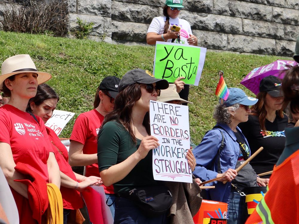 The image shows a group of people participating in a protest or rally, positioned in front of a stone wall and a grassy slope. The participants are holding signs with various messages. In the foreground, a woman wearing a green top and black cap holds a sign that reads, "THERE ARE NO BORDERS IN THE WORKERS' STRUGGLE." To her right, another person wears a blue jacket and a denim cap adorned with a small rainbow flag, holding drumsticks. Several individuals wear red T-shirts with a logo and text. The background features a person standing on the grassy slope, holding a sign with "ya Basta" in large letters. The participants display a diverse range of clothing and accessories, highlighting different messages and solidarity symbols.