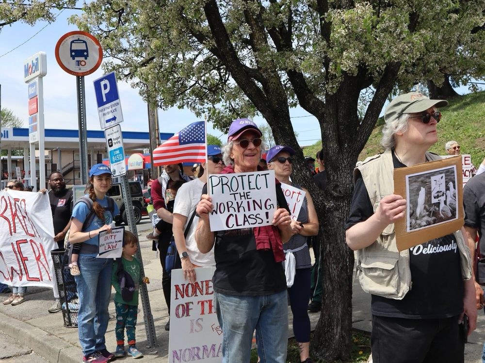 The image depicts a group of protesters standing on a sidewalk under a blossoming tree. A man in the foreground holds a sign reading "PROTECT THE WORKING CLASS!" while wearing a purple cap and sunglasses. To his right, another person carries a framed image of birds. Behind them, a woman with a blue cap and ponytail looks on, holding a sign. An American flag is visible amidst the crowd. Several other protest signs and banners with messages are partially visible. In the background, a Mobil gas station is seen, along with parking and bus stop signs.
