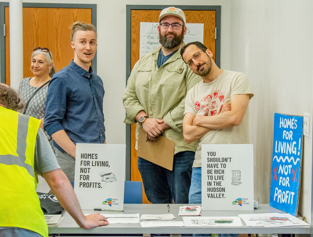 The image shows a group of five individuals standing behind a table displaying various signs and materials, likely at a community event or meeting. Three people in the center stand closely together, directly looking at the camera. They appear friendly and relaxed, with one person wearing a hat and holding a clipboard. Another person, to the left, is wearing a blue button-up shirt and is speaking, while another to the right in a printed t-shirt is slightly leaning forward. On the table are two white signs with bold text; one says, "HOMES FOR LIVING, NOT FOR PROFITS," and the other reads, "YOU SHOULDN'T HAVE TO BE RICH TO LIVE IN THE HUDSON VALLEY." Additional materials and flyers are spread across the table. To the far right, a blue sign with white and red lettering states, "HOMES FOR LIVING! NOT for PROFIT." More people are visible in the background on the left side of the image, including a person in a high-visibility vest, partially out of frame.