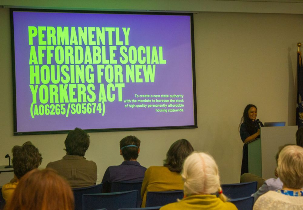 The image shows a presentation in a conference room. A woman stands at a podium on the right side, speaking into a microphone. She is smiling and appears to be engaging with the audience. A large screen to her left displays a purple slide with bold, bright green text covering most of its surface. The audience is seated in front of the screen, with the backs of their heads visible in the foreground. The room has a simple setup with blue chairs and a plain wall behind the screen. An American flag is partially visible on the right side of the image beside the presenter.