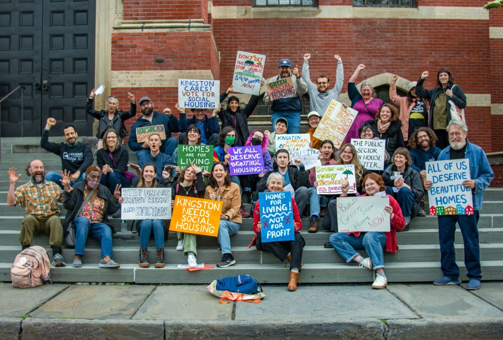 The image shows a group of about 25 people of varying ages gathered on the steps in front of a building with red and beige brickwork. They are smiling and holding a variety of signs advocating for social housing. The group is diverse, and they are showing enthusiasm with raised fists and animated expressions. The background includes a large, dark door and part of a window. The signs vary in color and content, displaying messages that support social housing and housing rights.
