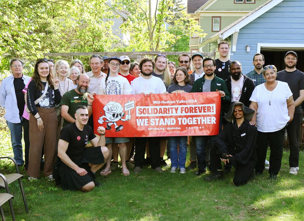 The image depicts a diverse group of people, approximately twenty, standing together outdoors in front of a blue house. They are holding a bright red banner with bold white text. The group is smiling and appears to be enjoying a sunny day. The background features greenery with trees and shrubs, adding to a pleasant, natural setting. The individuals are wearing casual clothing, and two people are kneeling in the front, each holding one end of the banner.