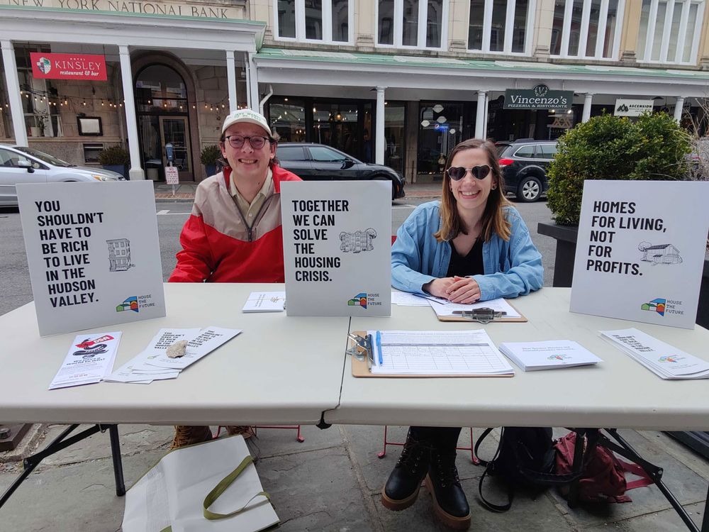 The image depicts a street scene with two people seated behind a rectangular table. The table is set up outside in front of a storefront and is covered with several items, including pamphlets, clipboards, and posters. The person on the left is wearing glasses, a red and beige jacket, and a hat. The person on the right is wearing sunglasses, a blue jacket, and a black shirt. Three signs are prominently displayed on the table, each containing different text related to housing issues. Behind them, there is a street with parked cars and a building with multiple signs including "New York National Bank" and various restaurants. There are also decorative lights hanging in the background.
