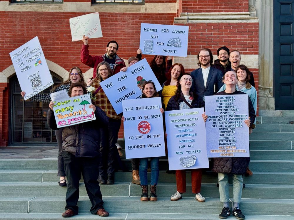 A group of fourteen people stand on the steps of a brick building holding various protest signs about housing. The individuals are diverse in terms of age and attire, smiling and appearing enthusiastic. The top row holds signs with messages like “TOGETHER! WE CAN SOLVE THE HOUSING CRISIS” and “BUILD GREEN SOCIAL HOUSING NOW!”. Middle row signs include “WE CAN SOLVE THE HOUSING” and “HOMES FOR LIVING NOT FOR PROFIT!”. Individuals in the front hold horizontally oriented signs stating “Cover as many tenants as possible!” and one listing various professions followed by "ALL OF US DESERVE BEAUTIFUL HOMES". The background features a red brick facade and a large dark door.