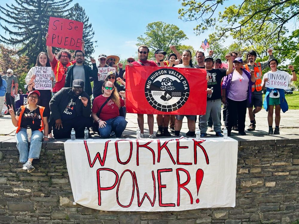 The image depicts a group of people participating in a rally or protest. The group consists of diverse individuals, some seated on a stone ledge and others standing behind them. They hold various signs and banners, including a prominent red banner reading "Mid Hudson Valley DSA" with a logo. Below them, a large white sheet with "WORKER POWER!" written in bold red letters hangs down the stone wall. Individuals hold signs with messages such as "Si Se Puede," "There Are No Borders On the Worker’s Struggle," "Worker Power," and "Union Strong." The scene is set outdoors on a sunny day with tall trees and a clear blue sky in the background. Some participants raise their fists, indicating solidarity.

