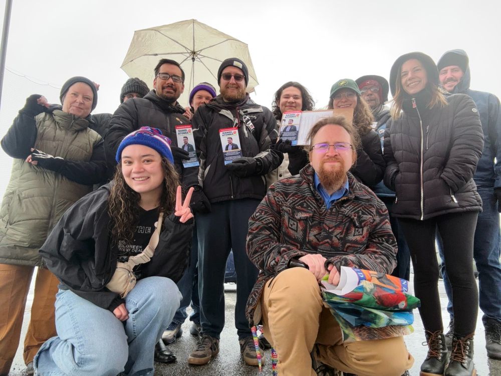 
The image shows a group of people gathered closely together. They are dressed warmly in winter clothing, with some wearing jackets, hats, and scarves. One person in the front is kneeling down, displaying a peace sign with a cheerful expression, while another person kneels beside them holding colorful papers. Behind them, individuals stand closely, some smiling, while at least two people hold up pamphlets featuring a man's image. An umbrella is visible above one person's head near the center of the group. The setting appears to be outdoors on an overcast day, suggested by the bright yet diffused lighting.