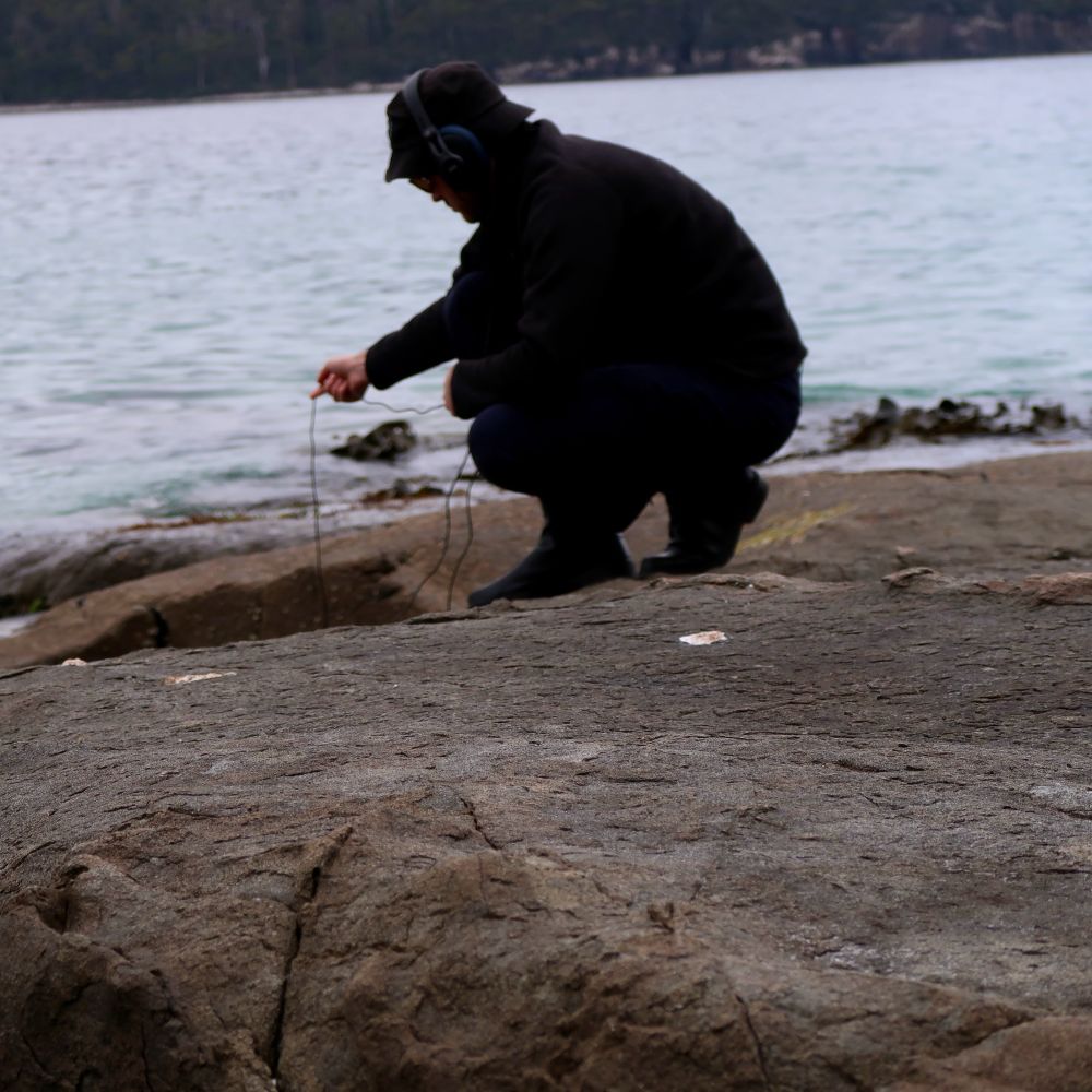 Photo of Spectrical crouching on a rock, dangling a microphone cable into the ocean