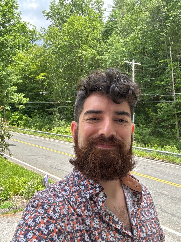 Selfie of John in front of a wooded area and a road. He’s wearing an orange, white, and blue floral collared shirt. He’s embraced his curly hair this year to go with his long reddish-brown beard. 