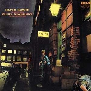 A blonde young man with a guitar has a foot raised onto a step on a landing under a sign K. West on nighttime street. David Bowie: The Rise and Fall of Ziggy Stardust and the Spiders From Mars.