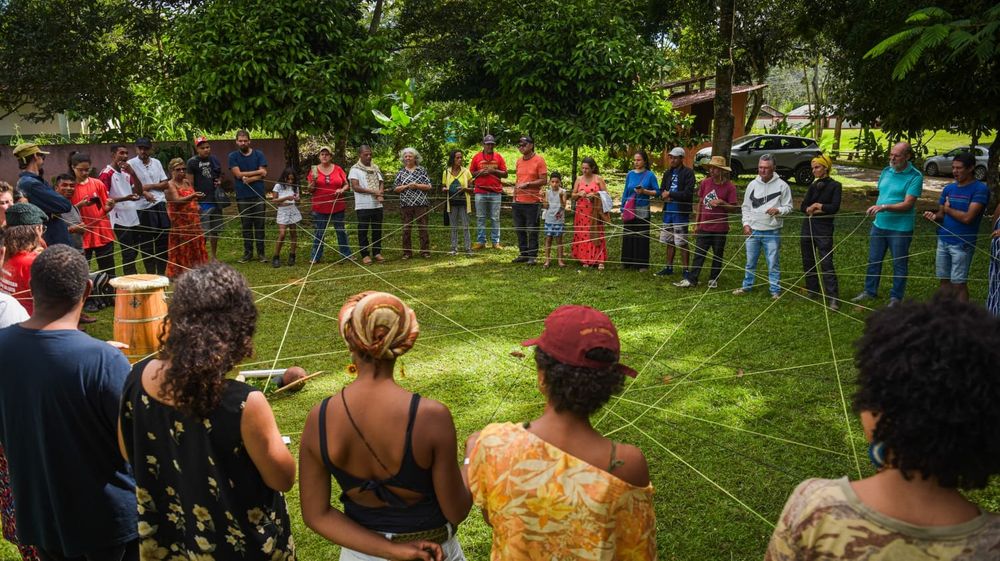 Grupo de cerca de 30 pessoas em círculo ao ar livre, em um gramado cercado por árvores. Cada pessoa segura um fio, formando uma grande teia de conexões no centro do círculo. No meio, instrumentos como atabaque e cabaças sugerem um momento de integração cultural ou espiritual. A atividade parece simbólica, representando união, interdependência e construção coletiva. A diversidade do grupo é visível, e o ambiente transmite acolhimento e conexão com a natureza.