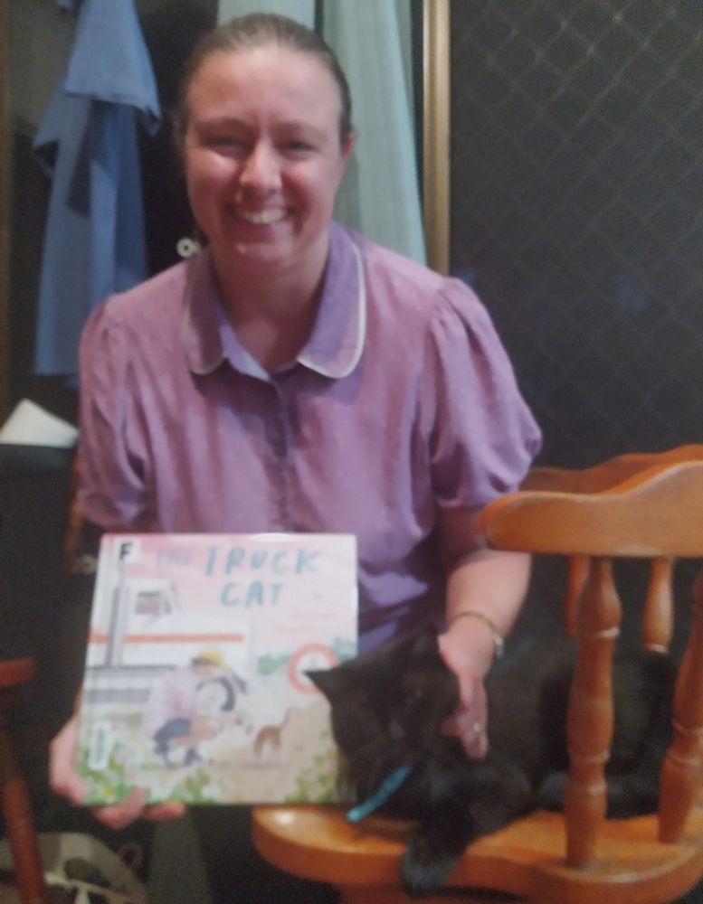 A smiling woman in a purple collared blouse holds a copy of the Truck Cat book whilst petting a black cat with a blue collar.