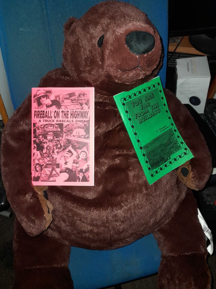 A djungleskog brown teddy bear holds 2 zines, the left is a red zine titled Fireball On The Highway: A Truck Rascals Zine and the right is a green zine titled You Are Far From The Building