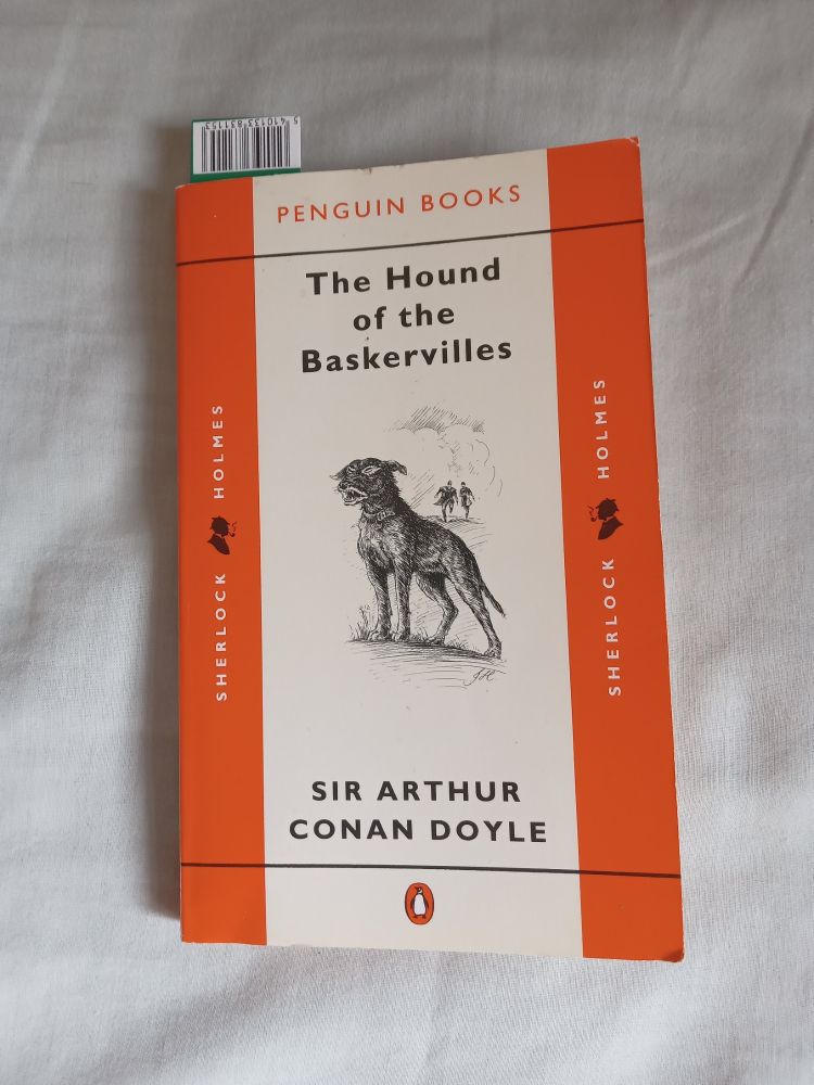 Front cover of a Penguin paperback of 'The Hound of the Baskervilles' by Arthur Conan Doyle. In middle is a drawing of a baying hound, with Holmes & Watson in the distance running towards it.