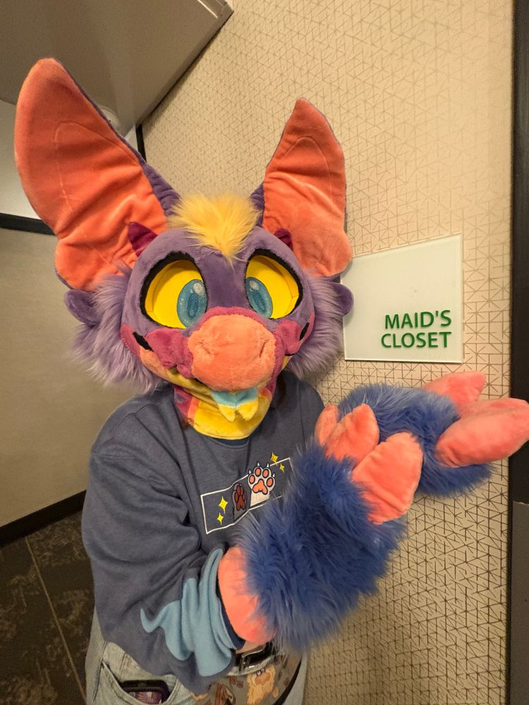 Photo of a person wearing a purple dragon fursuit, next to a hotel sign that says “maid’s closet”