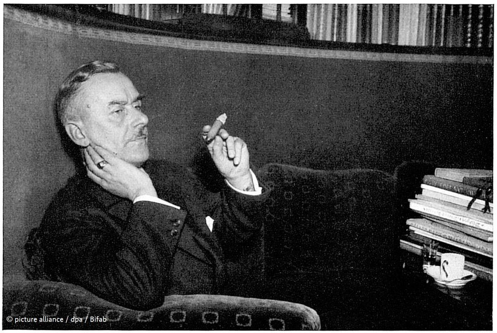 Thomas Mann auf seiner Couch vor einem Bücherregal sitzend, in der Hand hält er eine Zigarre.