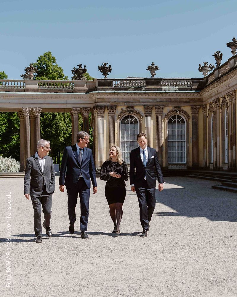 Bei einem Pressetermin am Schloss Sanssouci in Potsdam waren heute auch der Generaldirektor der Preußischen Stiftung Schlösser und Gärten Berlin-Brandenburg Christoph Martin Vogtherr, Berlins Kultursenatorin Sarah Wedl-Wilson und Georg Friedrich Prinz von Preußen dabei. Alle vier laufen gut gelaunt am Schloss entlang. Rechts sieht man die verzierte Fassade, links im Hintergrund einen Kolonadengang.