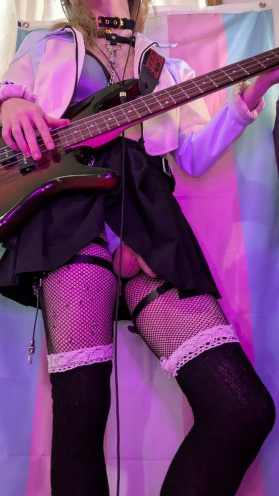 Tmiah standing in front of the camera and posing with their bass guitar. They're wearing plenty of black straps around their thighs and chest, a white cropped jacket, black thigh-highs, fishnet and skirt, a blue bra and matching panties, pulled to the side revealing their soft cock and balls from under the skirt.