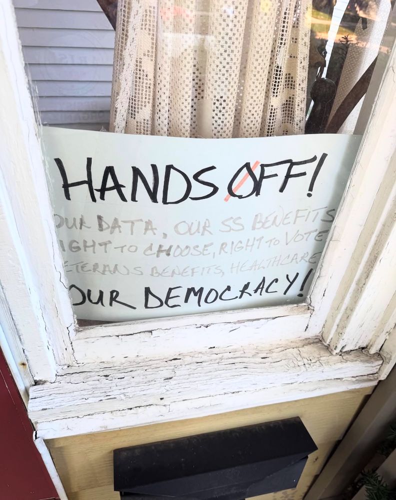 The photo features a window with a white painted border, and a sign placed in the glass that says "HANDS OFF: Our data, our social security benefits, right to choose, right to vote, veterans benefits, healthcare, OUR DEMOCRACY!"