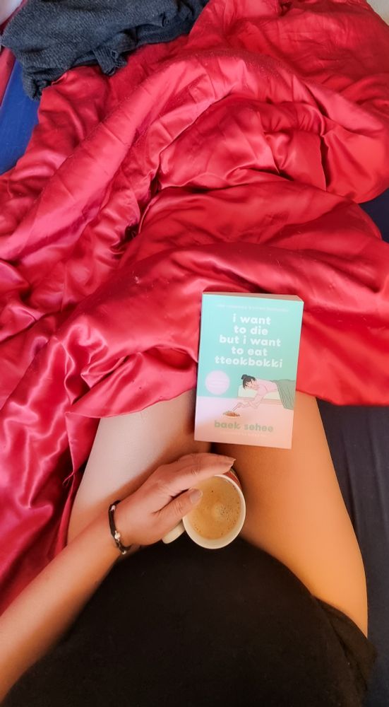 Kaffeetasse auf dem Schoß mit dem Tassenhenkel auf 8 Uhr (für meinen Lieblingsstatistiker) Schwarzes T-Shirt Ein Buch auf dem Schoß "I want to di* but I want to eat tteokbokki" Rote Satin Bettdecke Blauer Matratzenüberzug