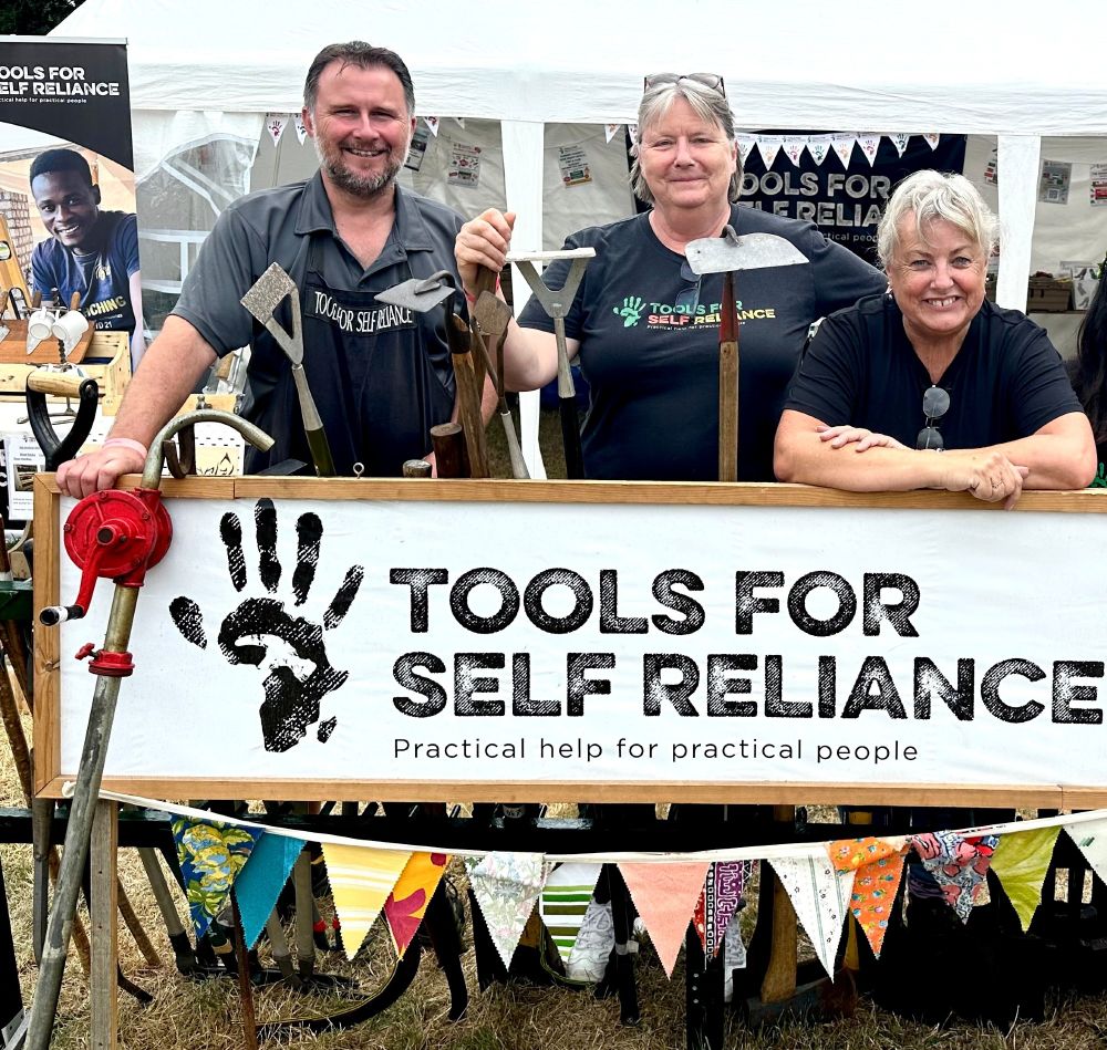 3 people standing behind a Tools for Self Reliance sign. There's some bunting and tools too.