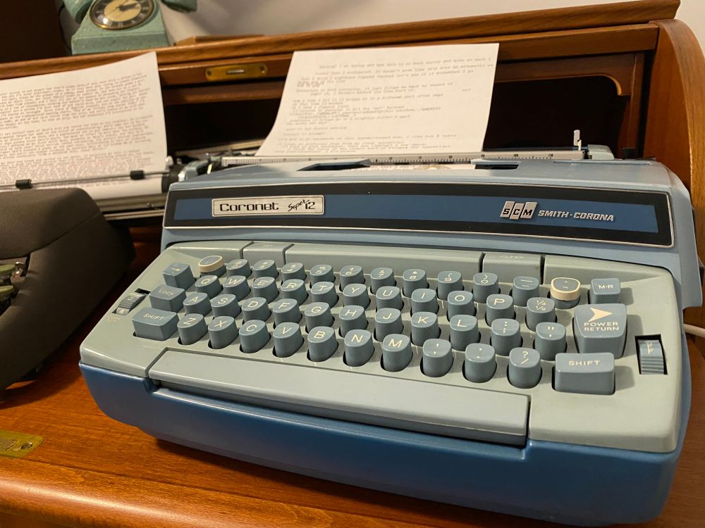 A baby blue Smith-Corona Coronet electric typewriter, sitting next to a still-beloved Smith-Corona manual typewriter