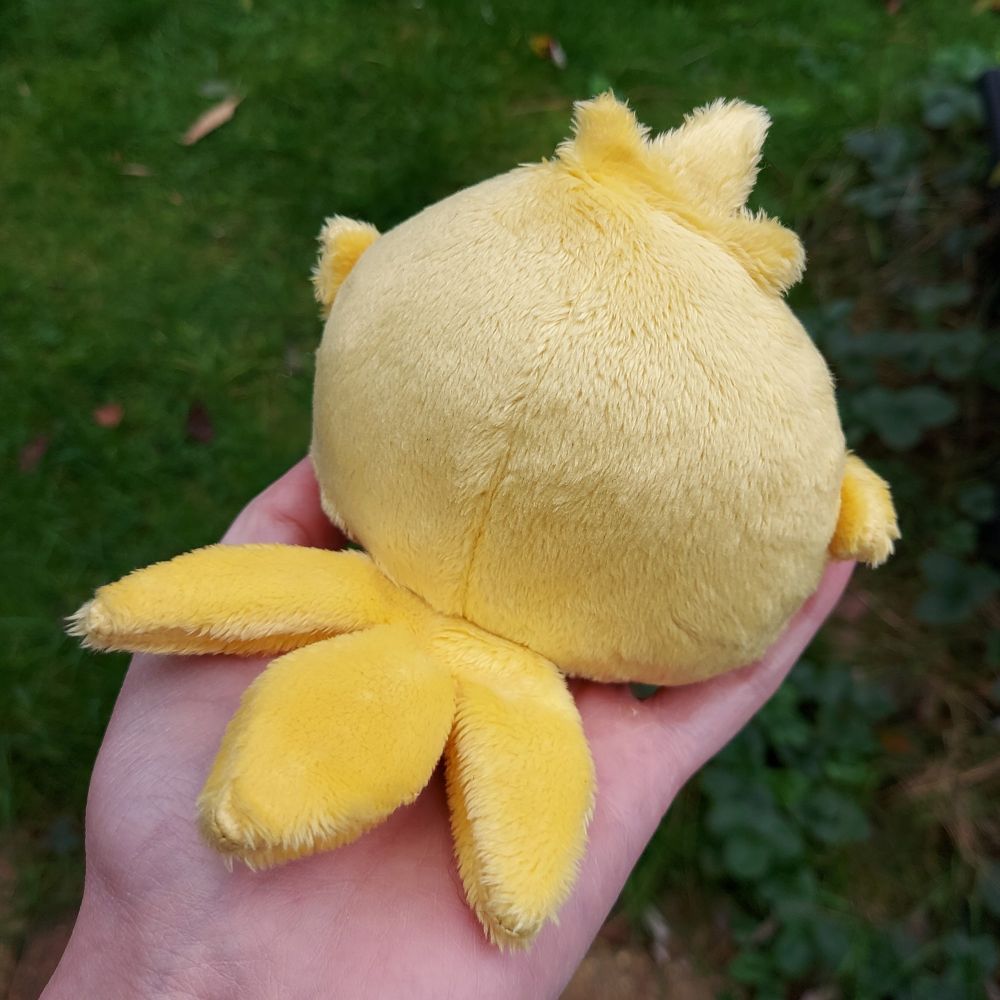 A photo of a hand holding a dumpling-shaped plush yellow chocobo chick. This is the back view of the plushie.