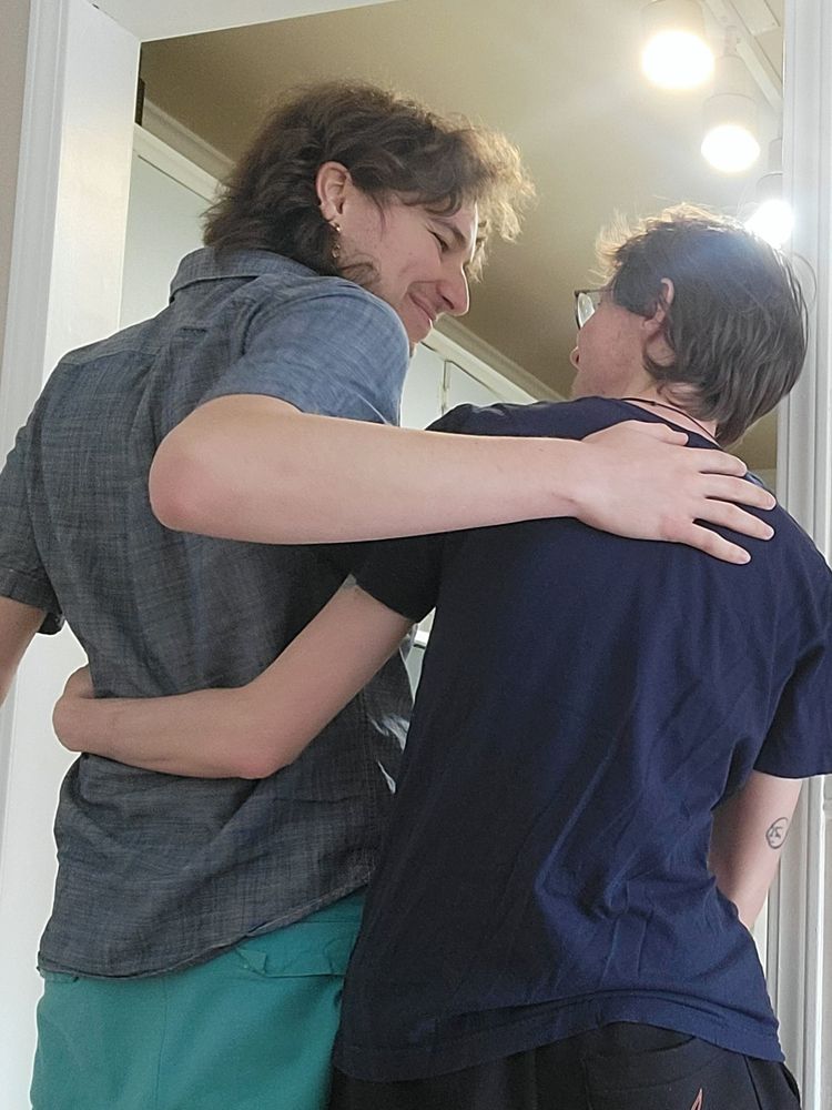 The back of one tall slim person smiling and hugging a short slim person. They are doing a pose to help the game's illustrator to draw one CG for the game. 