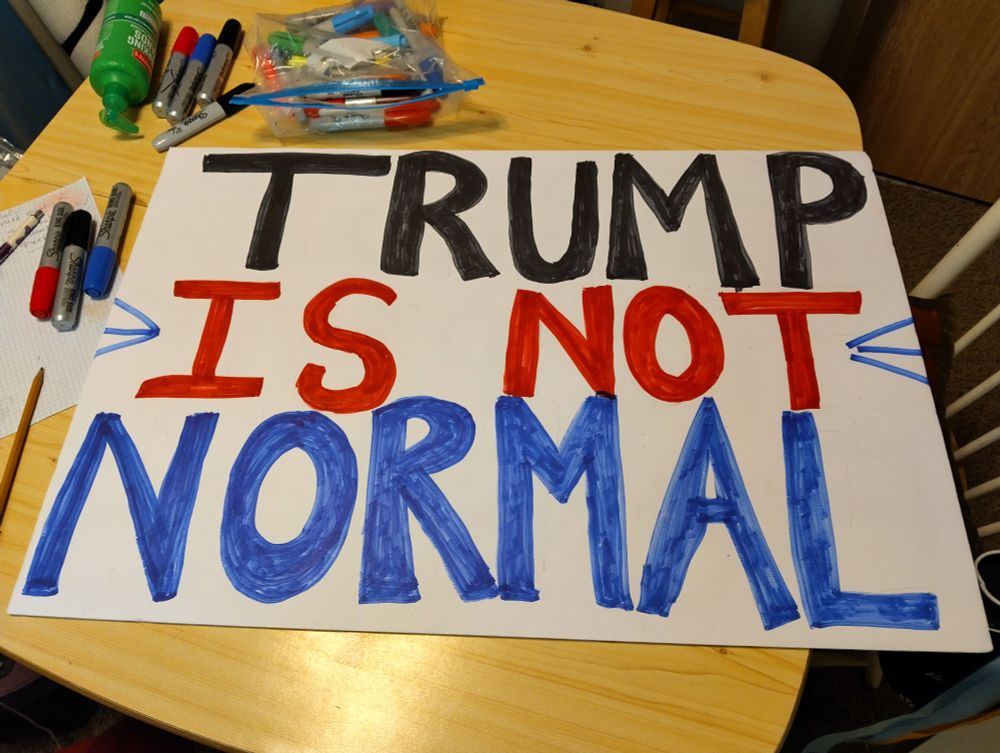 On a small table strewn with markers sits a handwritten sign waiting to spell.out the obvious to a cars driving by: TRUMP IS NOT NORMAL 