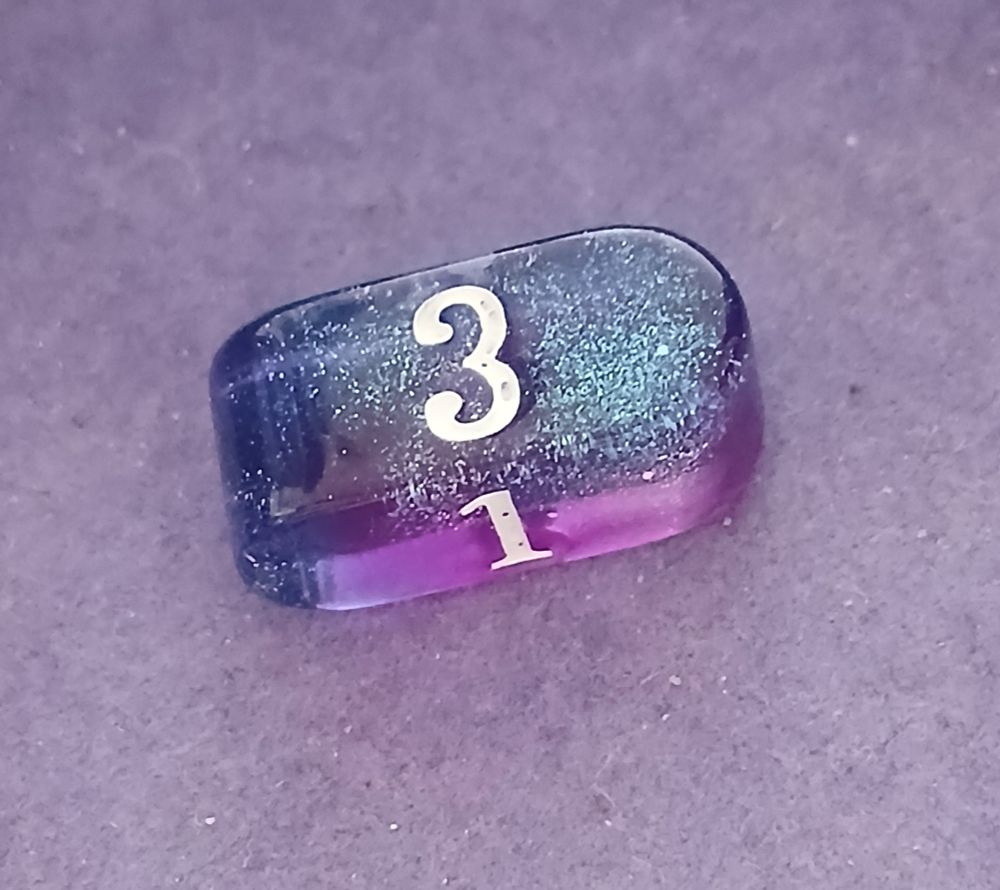 Sparkly purple 4-sided die in a rectangle shape with one side rounded in one direction and the other rounded in the other. 