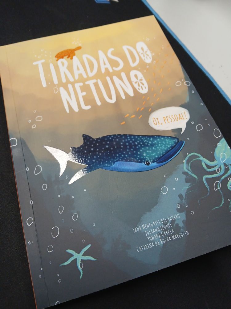 Descrição de imagem:
A foto, tirada por cima, mostra a capa de um livro infantil de capa mole. O livro tem o título "Tiradas do Netuno" escrito na parte superior com uma fonte branca e arredondada. A ilustração na capa mostra um cenário do fundo do mar. No centro, um tubarão-baleia azul-escuro com pintas brancas e um grande sorriso. Ele está de frente, com uma pequena bolha de fala que diz "Oi, pessoal!". No fundo, em tons de laranja e marrom, uma tartaruga-marinha nada na direção do título. Mais abaixo, em tons de azul, há um polvo e uma estrela-do-mar. Os nomes dos autores e ilustradores estão na parte inferior direita da capa. O livro está sobre uma superfície escura.
