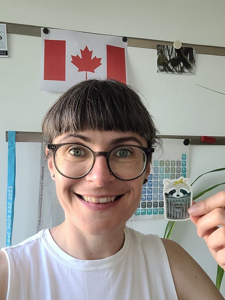 Stephanie Jones standing in front of printed Canadian flag holding a sticker of a cartoon Toronto racoon in a bin! 